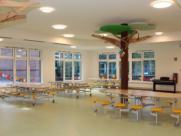 Lichtdurchflutete Mensa mit vielen Tischen und Stühlen und Holzbaum und Piano in der offenen Ganztagsschule Harksheide Nord in Norderstedt